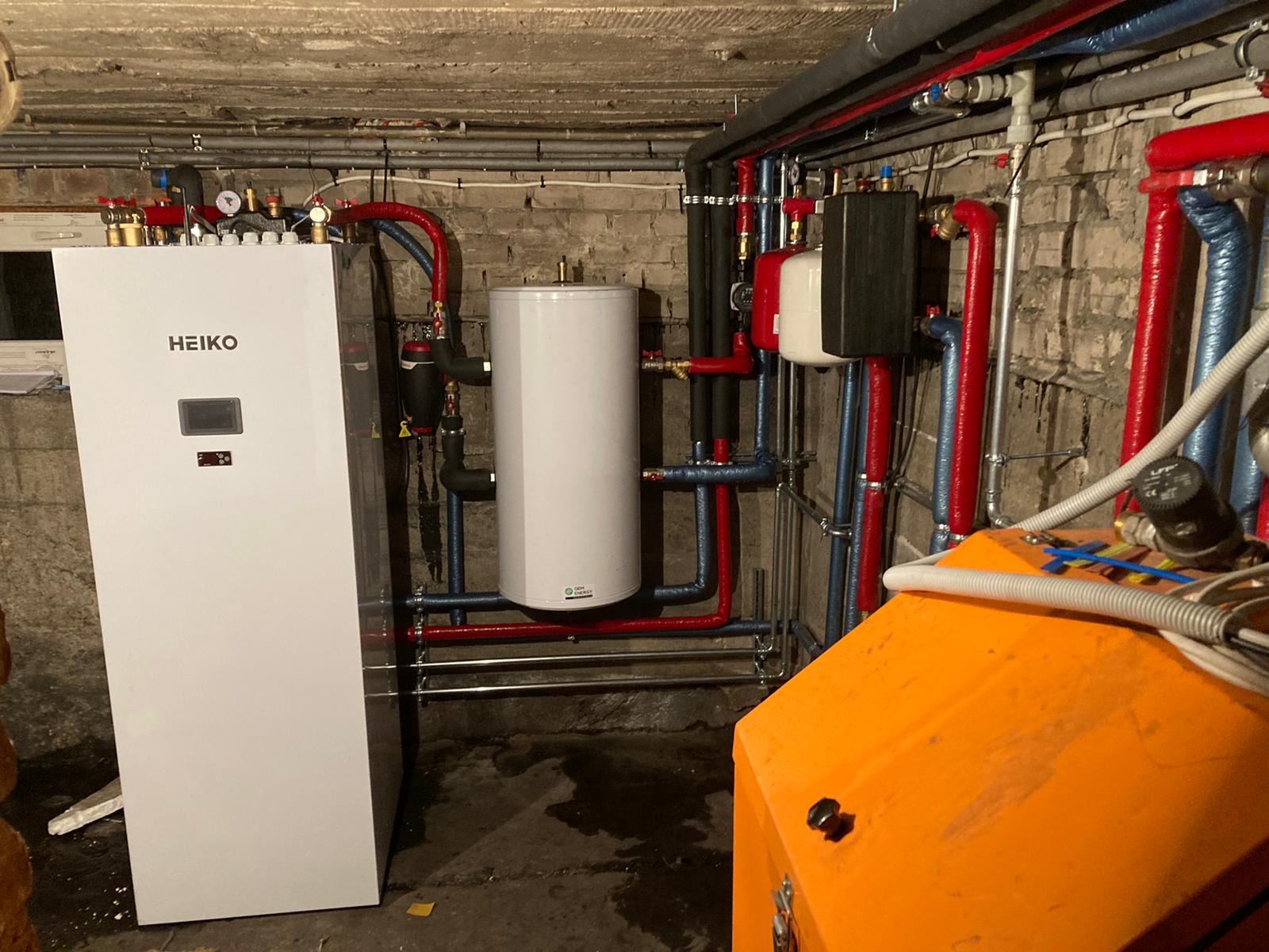 Instalacja pompy ciepła HEIKO z białym zbiornikiem buforowym, widoczne rury w kolorach czerwonym i niebieskim, instalacja w piwnicy z surowym betonowym sufitem i ceglanymi ścianami, pomarańczowa...
