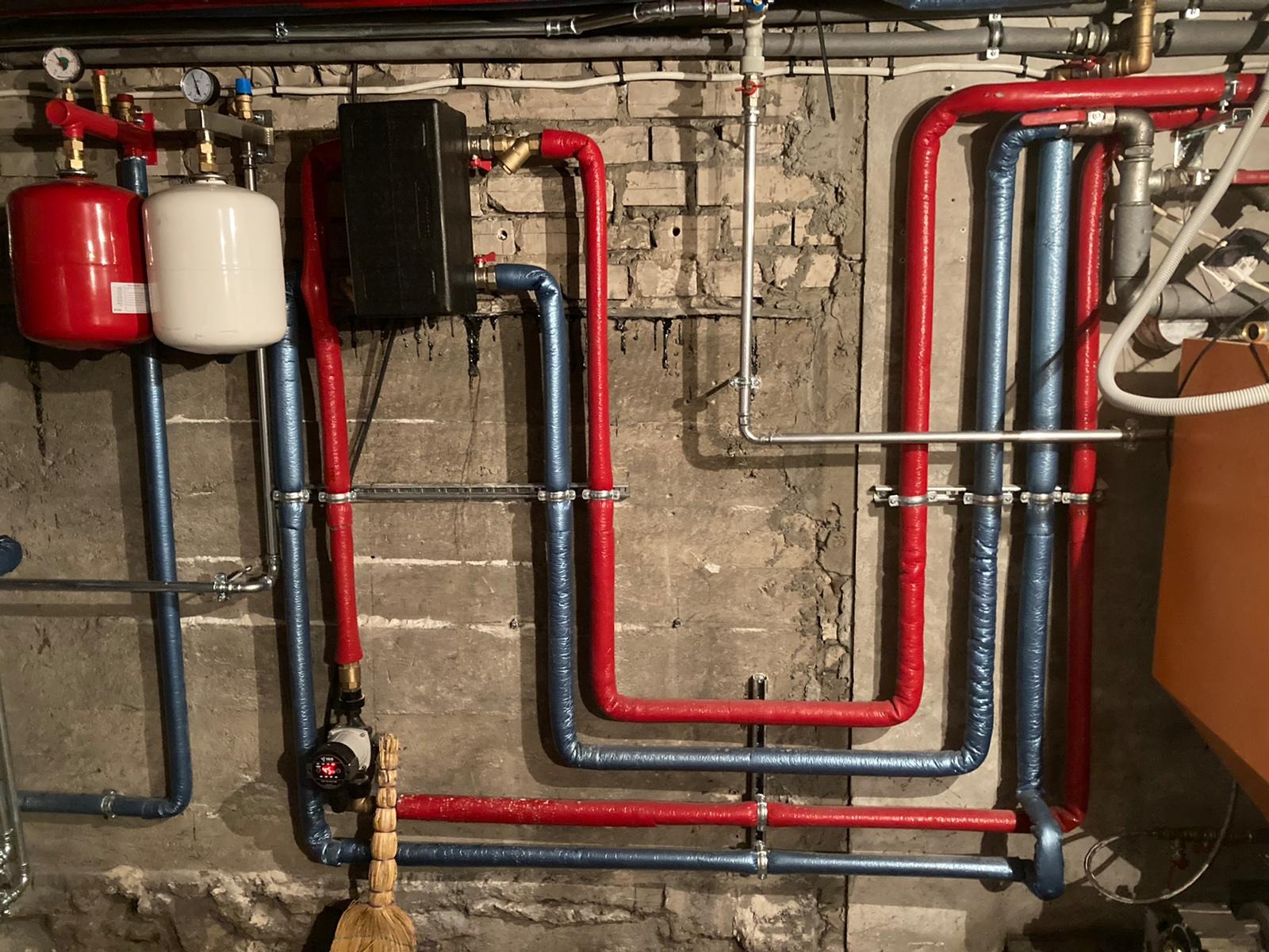 Instalacja grzewcza z czerwono-niebieskimi rurami izolowanymi termicznie na tle surowej ściany z cegieł i betonu, widoczny czarny zbiornik oraz pompa obiegowa z miotłą w rogu.