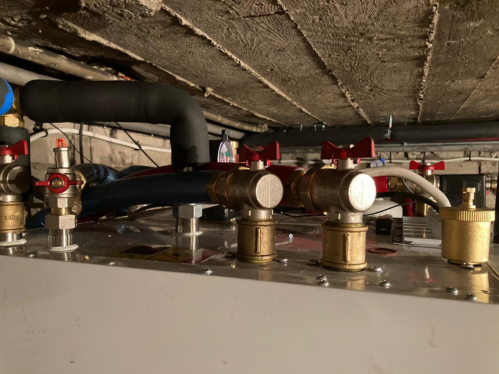 Szczegółowe ujęcie instalacji grzewczej z zaworami z czerwonymi pokrętłami, rurami w izolacji i surowym, betonowym stropem w tle.