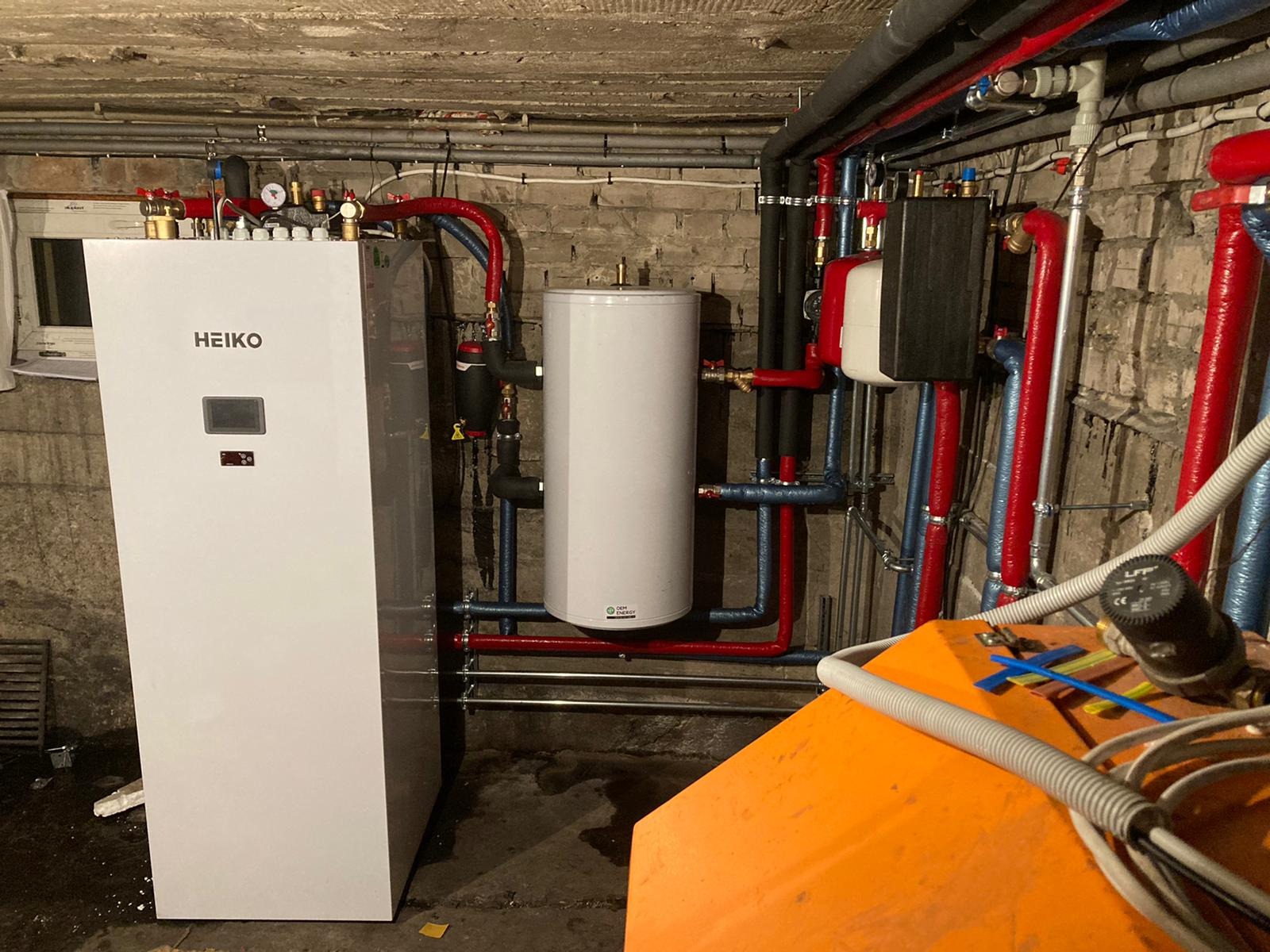 Biała pompa ciepła marki Heiko w piwnicy, z widocznymi rurami w izolacji czerwonej i niebieskiej, oraz białym zbiornikiem akumulacyjnym, na tle surowej ściany z cegły i betonowego sufitu.