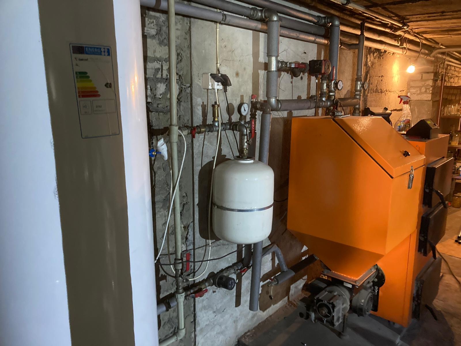 Pomarańczowy piec grzewczy na paliwo stałe z białym zbiornikiem wyrównawczym i instalacją hydrauliczną w kotłowni z surowymi ścianami.