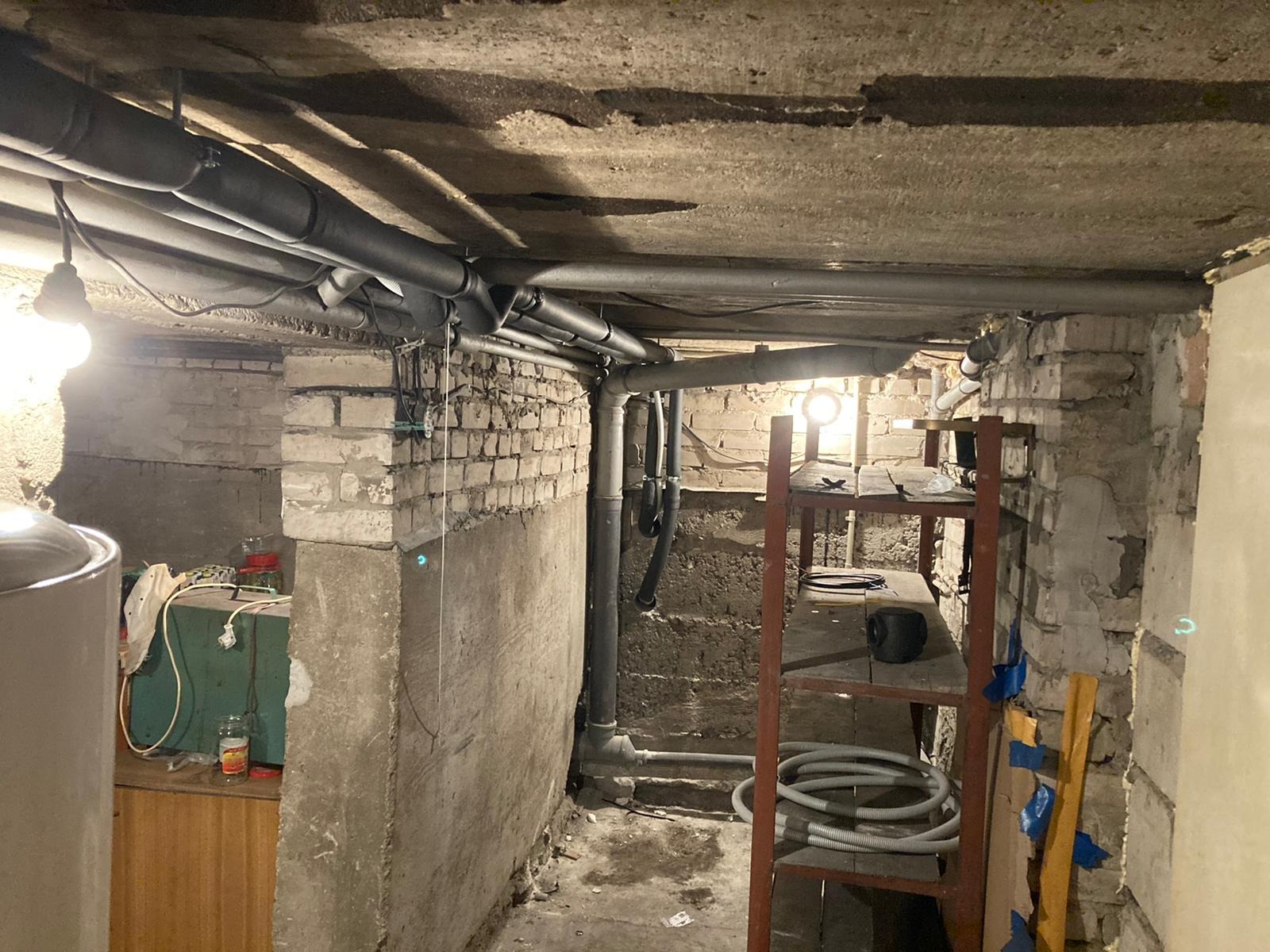 Rury instalacyjne, prawdopodobnie grzewcze, biegnące pod sufitem piwnicy z ceglanymi ścianami i prowizorycznym regałem.