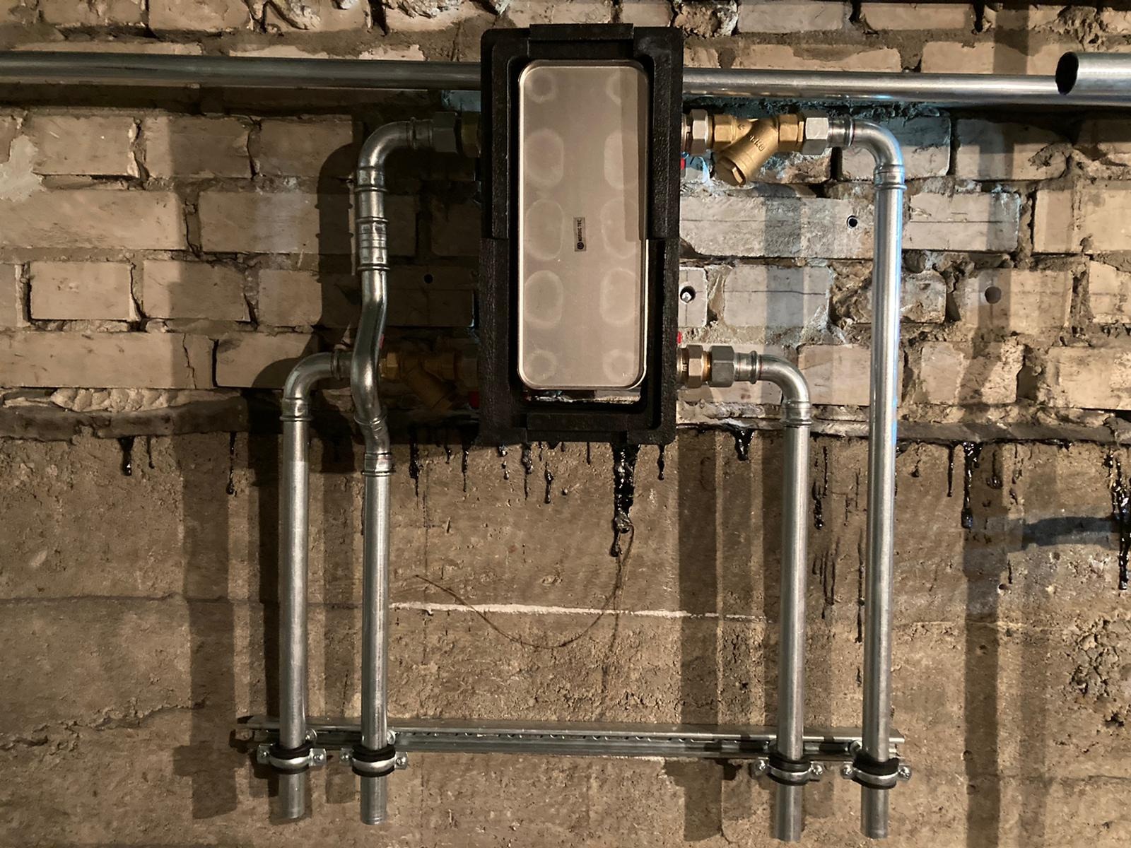 Szare rury instalacyjne z filtrem i czarną obudową urządzenia na tle ściany z cegły i betonu z zaciekami.