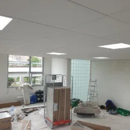 Montaż sufitu podwieszanego w biurze: widoczny stelaż, płyty w kartonie na wózku transportowym, drabina i narzędzia na podłodze przygotowanej do montażu.