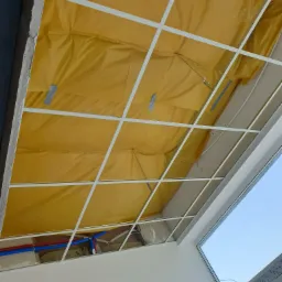 Montaż sufitu podwieszanego: widok na konstrukcję metalową, żółtą folię izolacyjną i instalację hydrauliczną w przestrzeni między sufitem a konstrukcją, z fragmentem okna w tle.