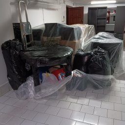 Brum brum Przeprowadzki - Meble i przedmioty zapakowane w folię i czarne worki, przygotowane do transportu, widoczne schody metalowe i fragment podłogi z białych płytek.