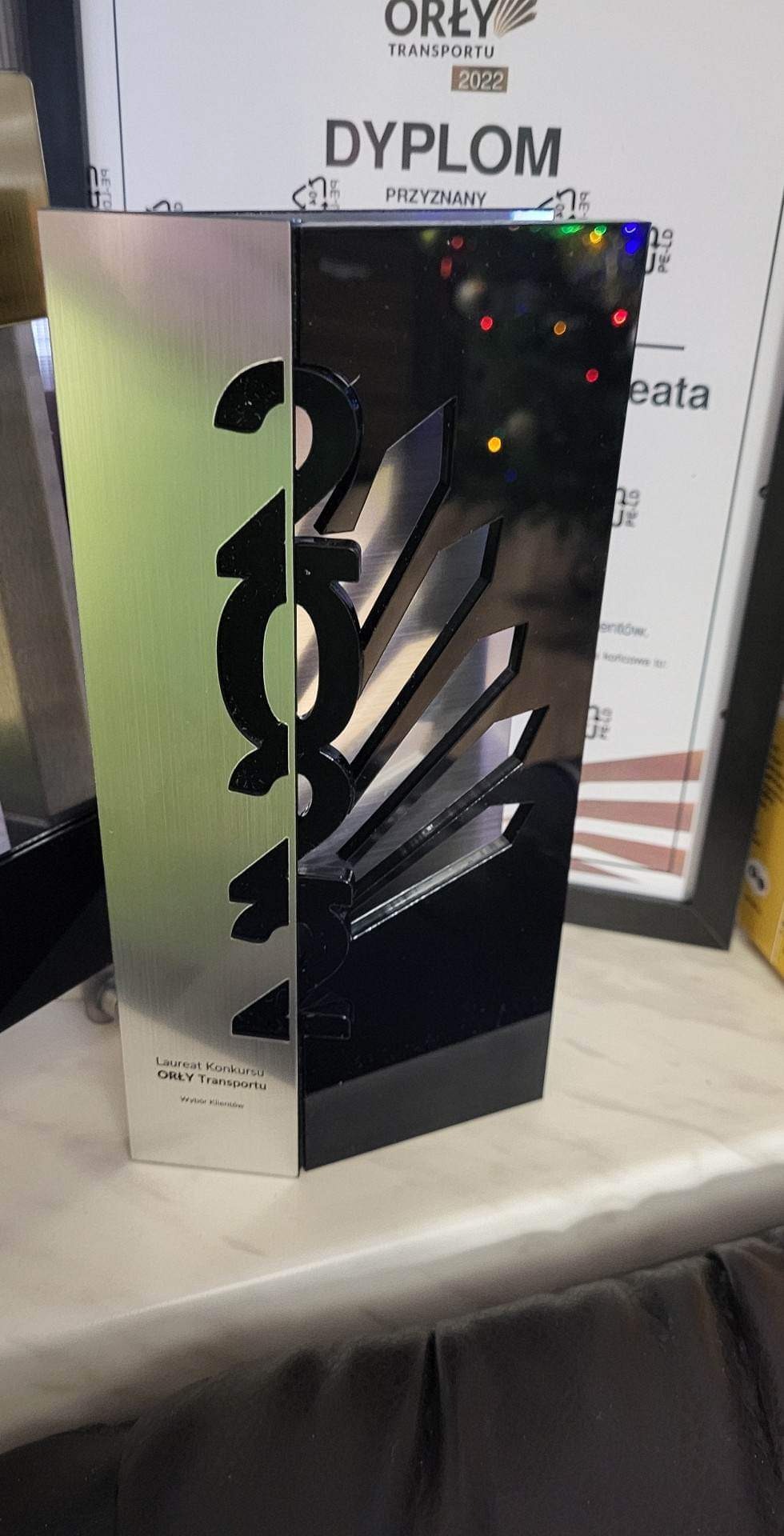 Statuetka konkursu Orły Transportu 2022, dwukolorowa (srebrna i czarna), z wyciętym napisem 2022, obok dyplom z nagrodą.