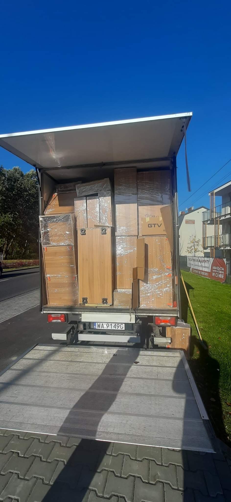 Załadunek mebli i kartonów owiniętych folią do otwartej skrzyni samochodu dostawczego z widoczną tablicą rejestracyjną WA 9149G na tle błękitnego nieba i zabudowań.