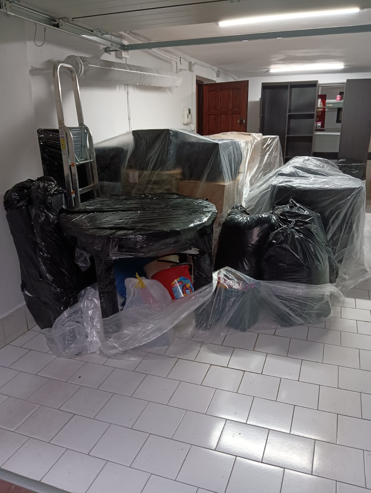 Meble i przedmioty zapakowane w folię i czarne worki, przygotowane do transportu, widoczne schody metalowe i fragment podłogi z białych płytek.