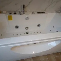 Biała wanna z hydromasażem, widoczne dysze wodne, bateria i marmurowe wykończenie ściany, brązowe płytki podłogowe.