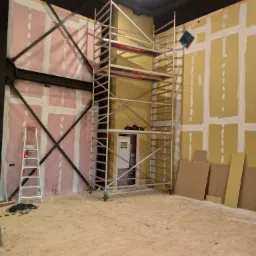 Wnętrze w trakcie prac przygotowawczych do wykończenia ścian; widoczne płyty kartonowo-gipsowe z nałożoną wstępną warstwą gładzi, metalowe rusztowanie, drabina, oraz panele podłogowe czekające...