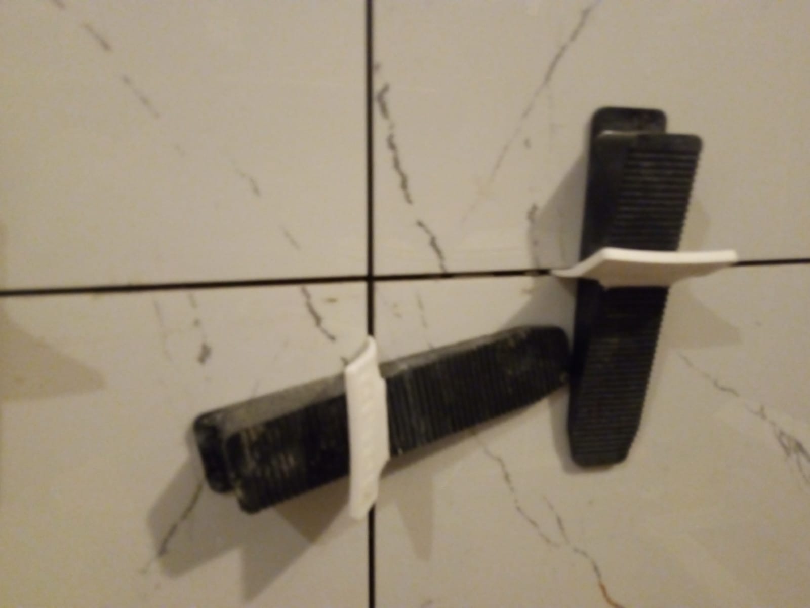 Dwa kliny dystansowe z białymi wkładkami, używane do poziomowania płytek ceramicznych na ścianie z jasnego marmuru.