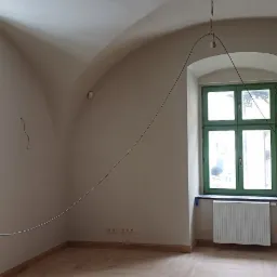 Wnętrze pokoju po remoncie z jasną podłogą ułożoną w jodełkę, świeżo pomalowane ściany w odcieniach beżu, sklepiony sufit, zielone okno i wiszący kabel od lampy.