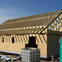 Konstrukcja drewniana domu w trakcie budowy, widoczna więźba dachowa i ściany z płyt OSB, na pierwszym planie zbiornik na wodę i toaleta przenośna.