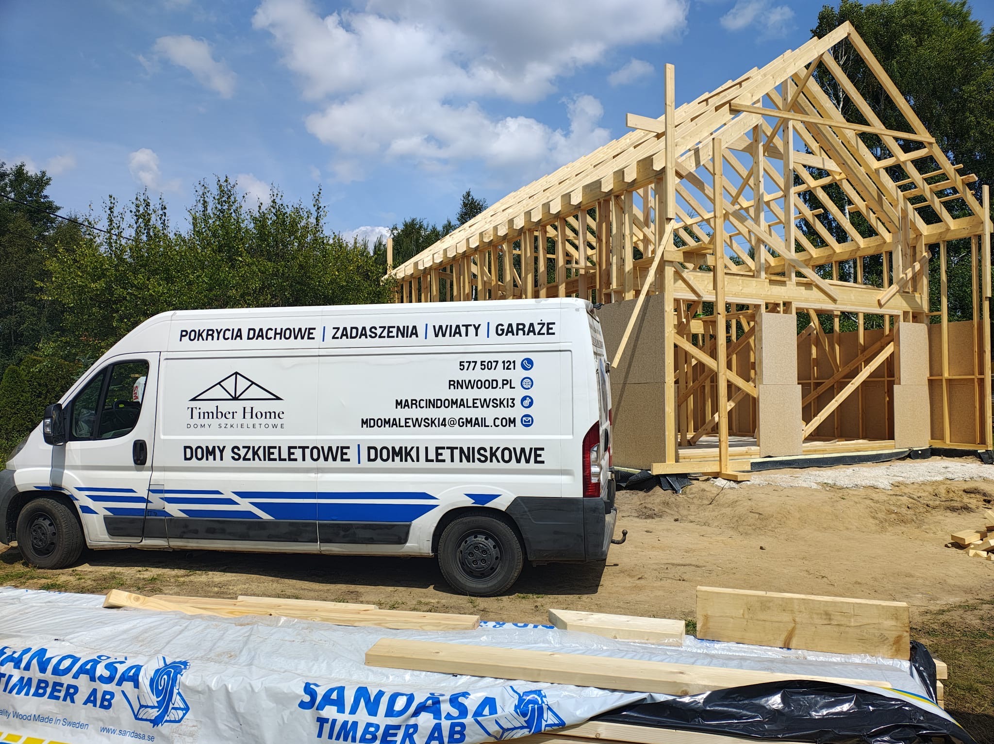 Konstrukcja szkieletowa domu w budowie, widoczna na tle firmowego busa Timber Home z danymi kontaktowymi. Częściowo zakryte folią z logo Sandasa Timber AB.