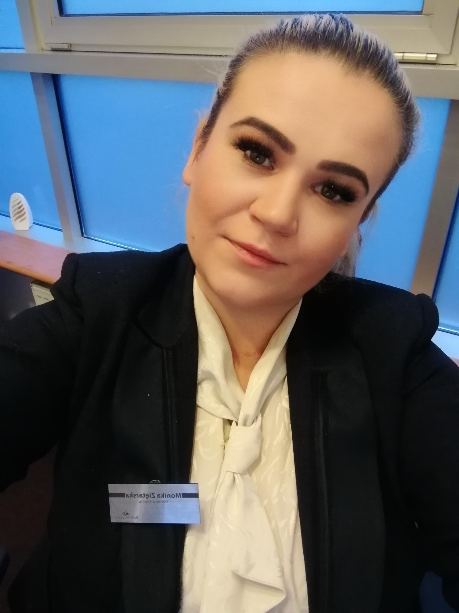 Portret kobiety w czarnej marynarce i białej bluzce z plakietką imienną, wykonany w biurze z oknem w tle.