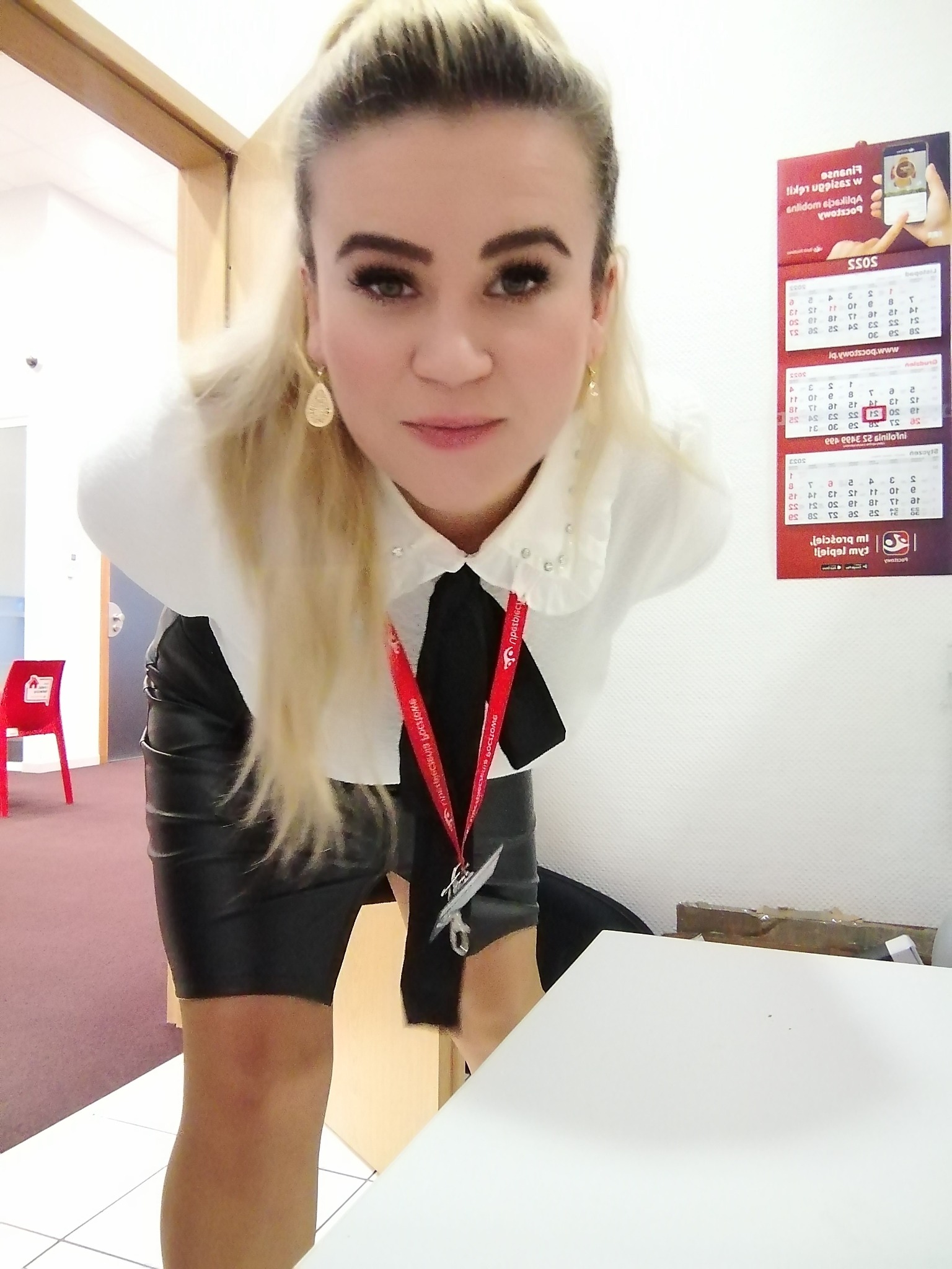 Pracownica banku z identyfikatorem na smyczy, pochylona nad biurkiem, w tle kalendarz bankowy i czerwone krzesło.