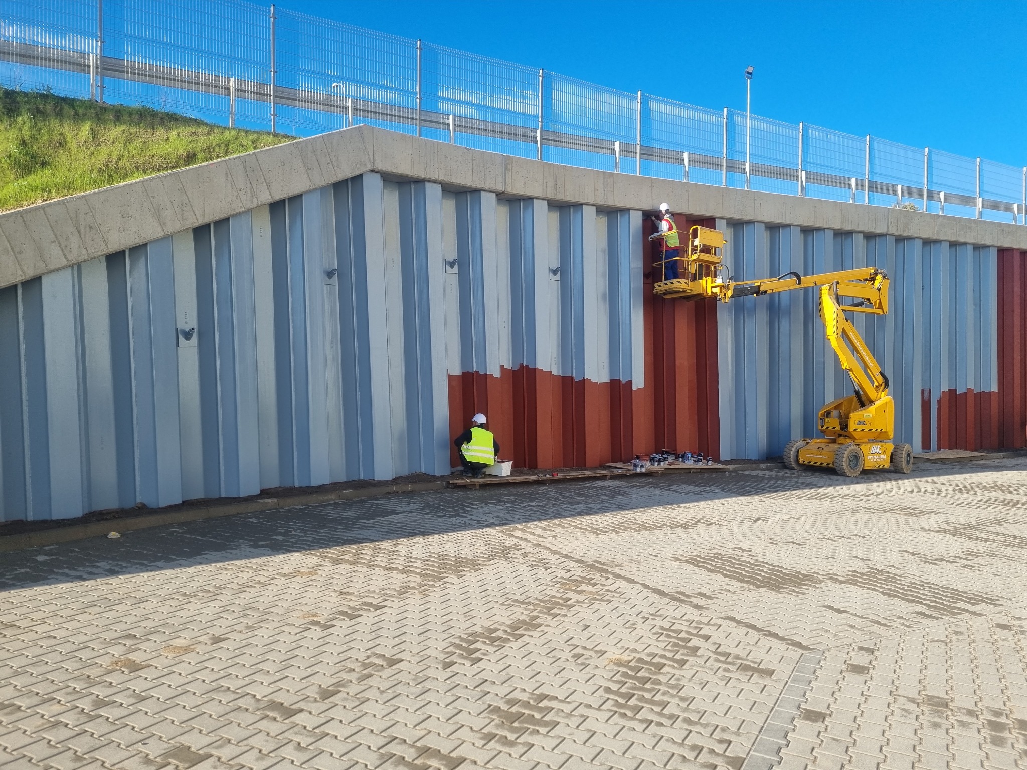Pracownik na podnośniku koszowym maluje na brązowo metalową ścianę wiaduktu, drugi pracownik siedzi na ziemi obok puszek z farbą.