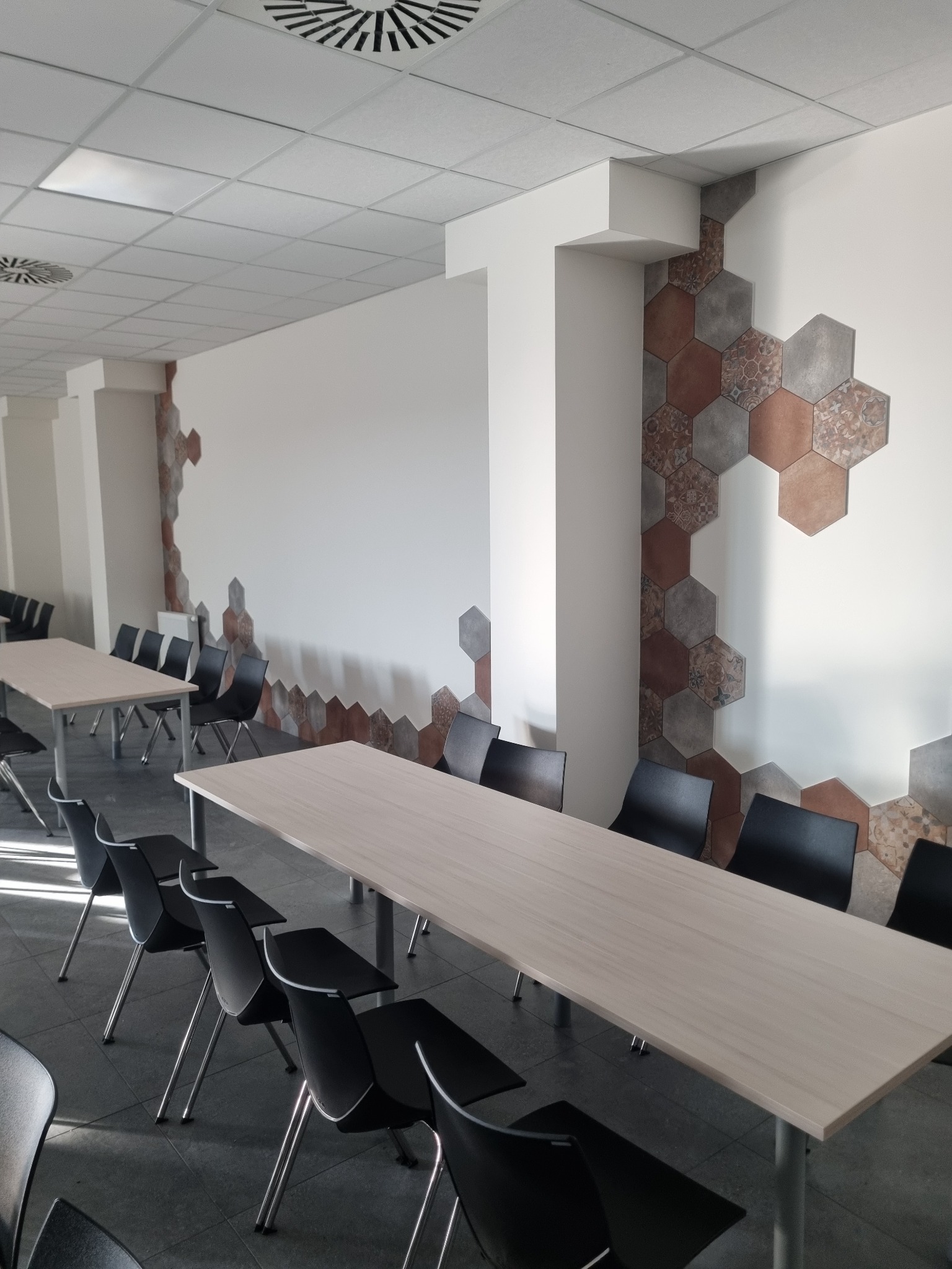 Sala konferencyjna z jasnymi stołami i czarnymi krzesłami, ściana ozdobiona heksagonalnymi płytkami w odcieniach brązu i szarości, tworzącymi nieregularny wzór.