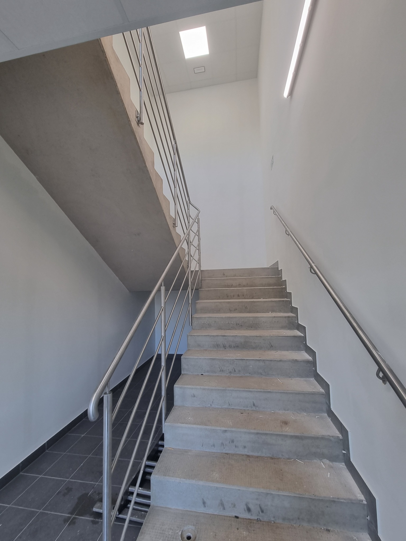 Klatka schodowa w biurowcu z betonowymi schodami i metalowymi poręczami, ściany pomalowane na biało, widok z dołu.