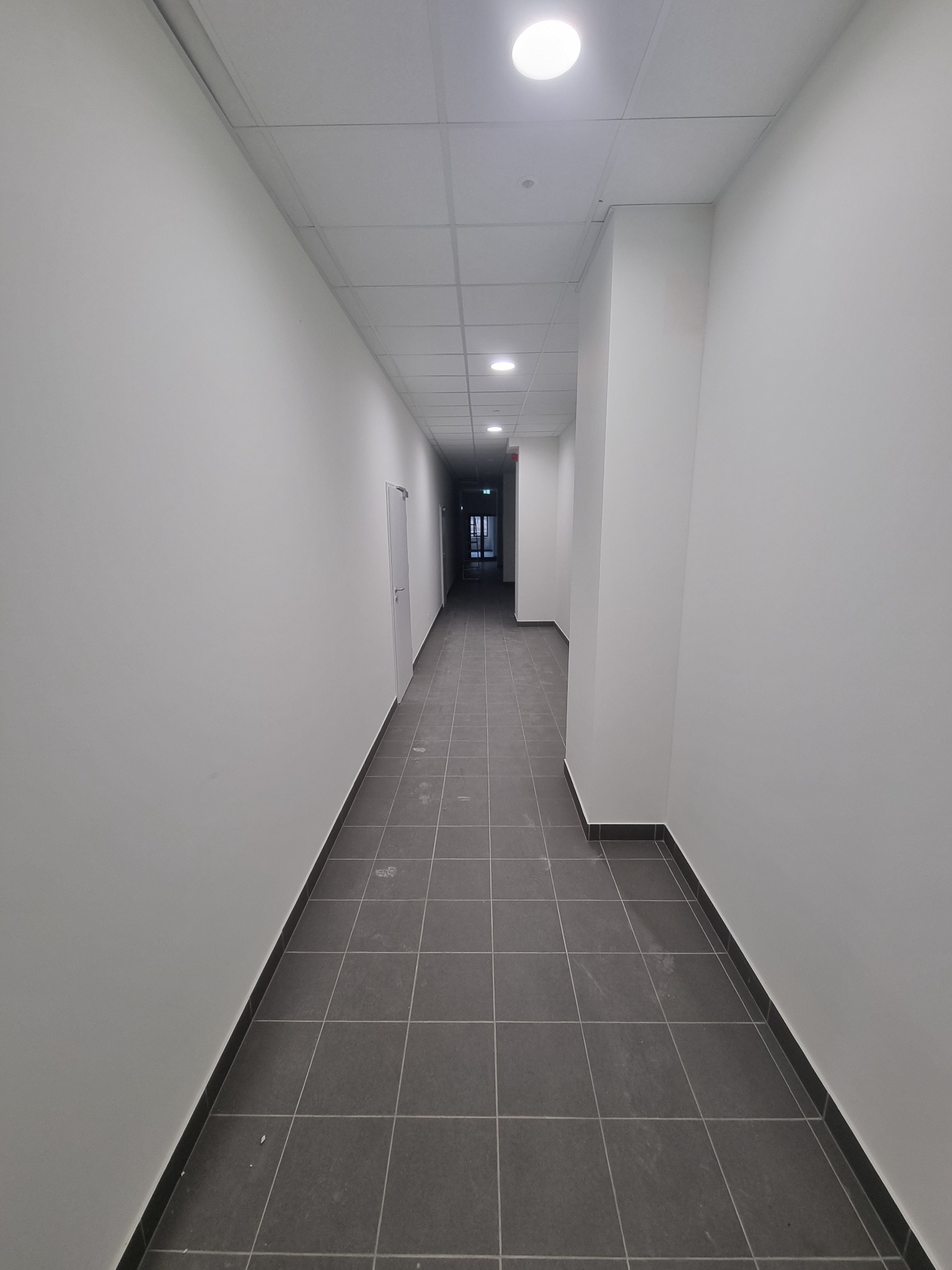 Długi, pusty korytarz biurowy z szarymi płytkami podłogowymi i białymi ścianami, oświetlony przez okrągłe lampy sufitowe, widoczne ślady po pracach wykończeniowych na podłodze.
