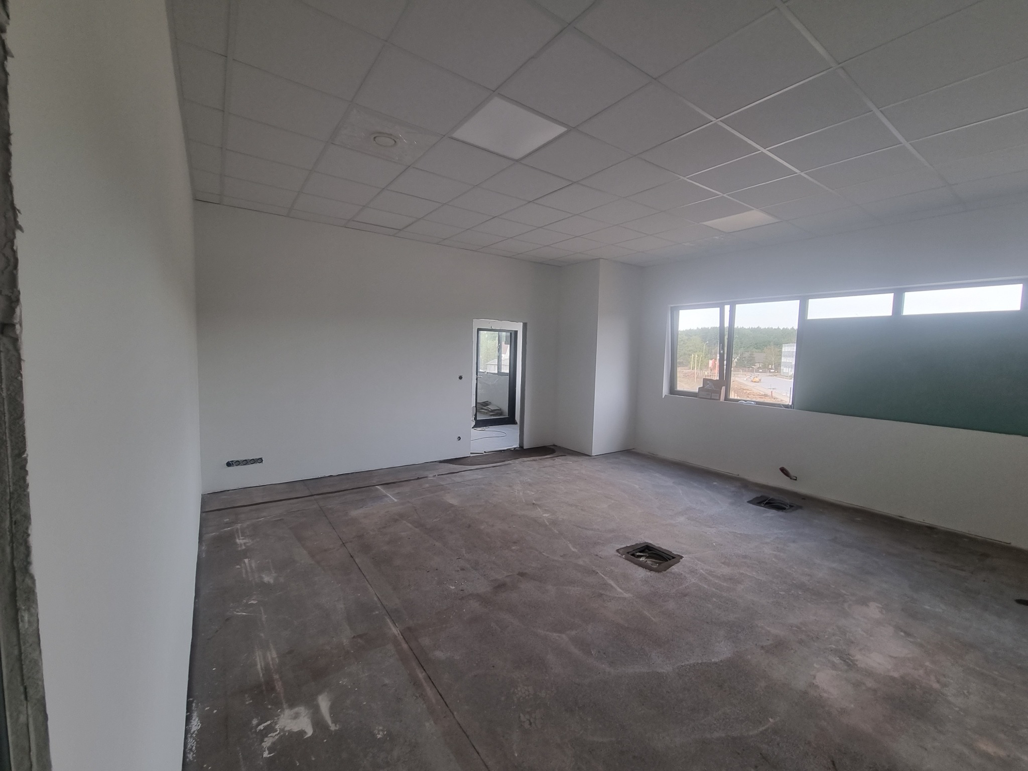 Pusty, jasny pokój biurowy w stanie surowym, z białymi ścianami po świeżym pomalowaniu, betonową podłogą przygotowaną do wykończenia, oknami z widokiem na zewnątrz i kasetonowym sufitem.