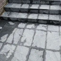 Schody zewnętrzne wyłożone szarymi płytkami imitującymi kamień, z widocznymi fugami i ceglanym fragmentem ściany po lewej stronie.