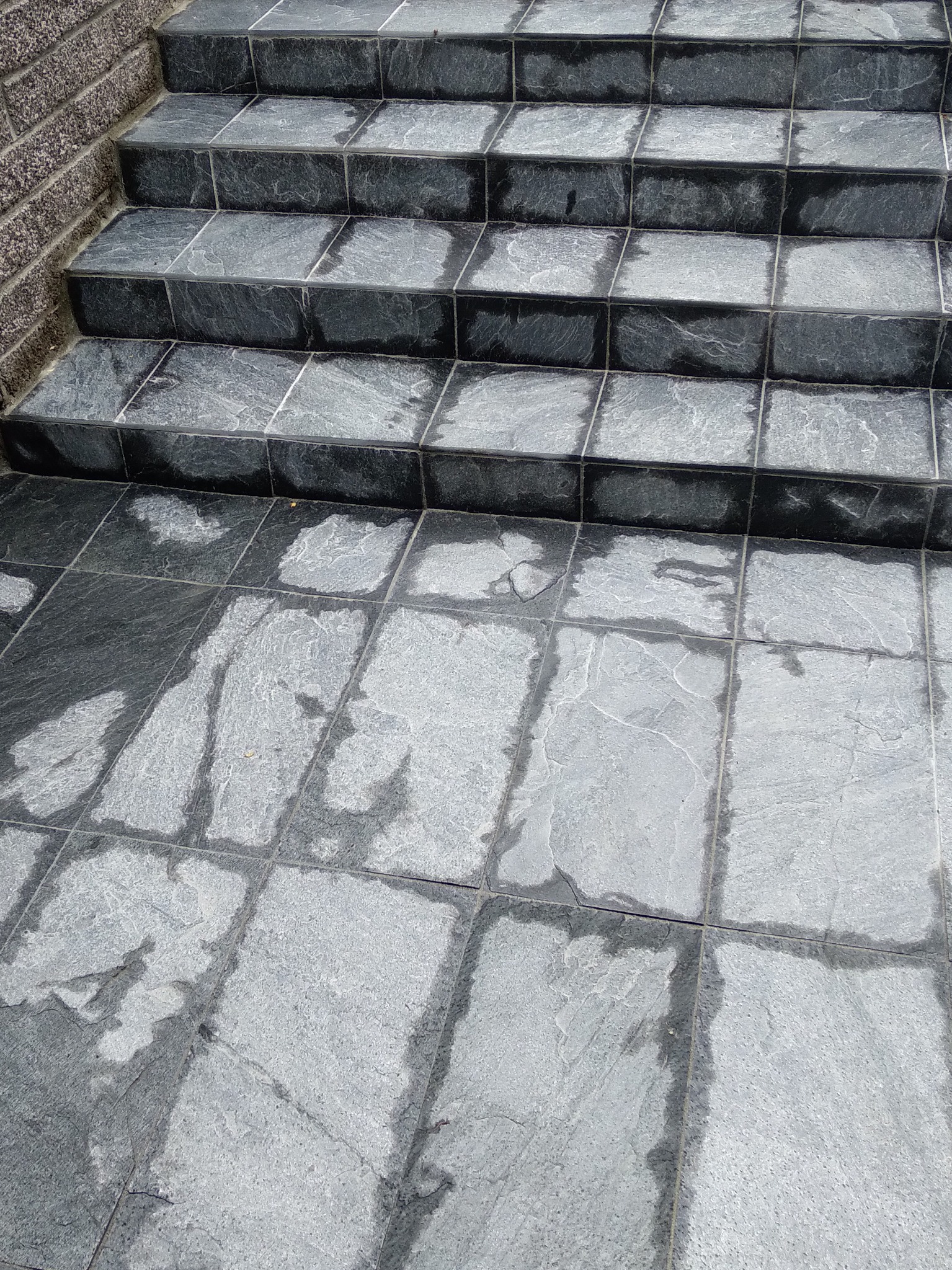 Schody zewnętrzne wyłożone szarymi płytkami imitującymi kamień, z widocznymi fugami i ceglanym fragmentem ściany po lewej stronie.