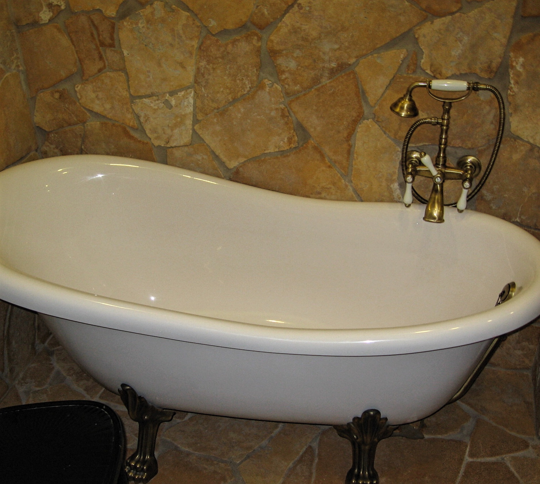 Wolnostojąca wanna na ozdobnych nóżkach z mosiężną armaturą i prysznicem na tle ściany wyłożonej nieregularnymi kamieniami w odcieniach beżu i brązu.