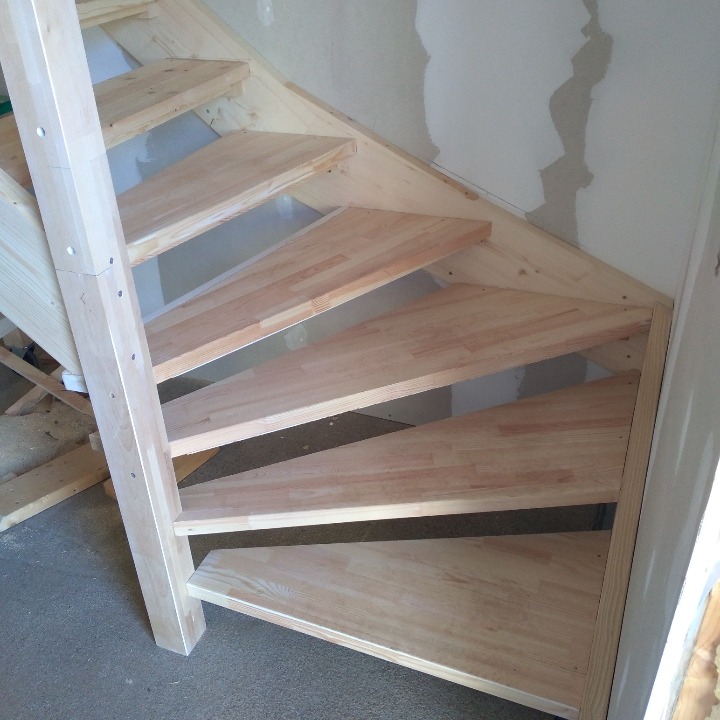 Drewniane schody z jasnego drewna w trakcie montażu, widoczne niedoskonałości na ścianie i surowy beton na podłodze.