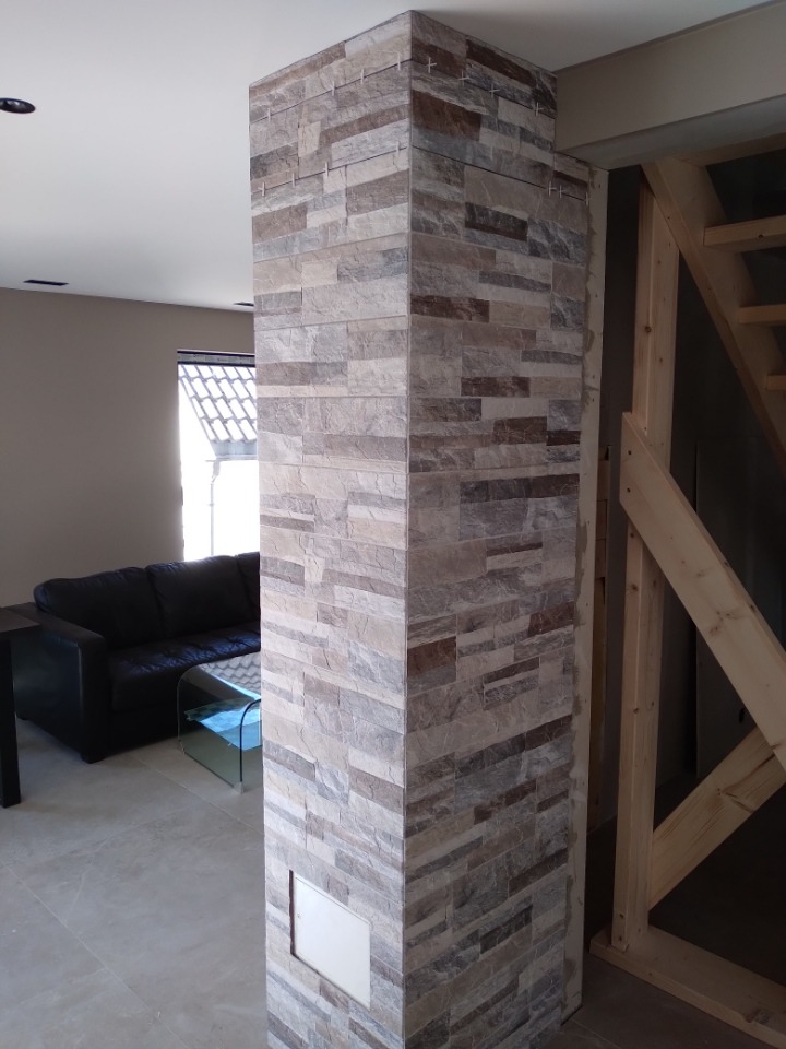 Kolumna wykończona dekoracyjną okładziną kamienną w salonie, widoczna klapka rewizyjna na dole kolumny, w tle czarna sofa i szklany stolik kawowy oraz drewniana konstrukcja schodów.