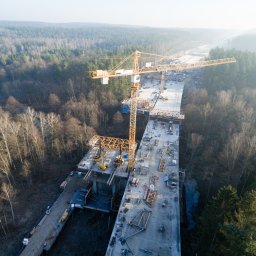 Geo-Control Tobiasz Gałazka - Zdjęcie z drona: budowa mostu w otoczeniu lasu. Dwa żółte dźwigi na betonowej konstrukcji. Widoczni pracownicy w pomarańczowych ubraniach. Leśne tło w lekkiej mgle.