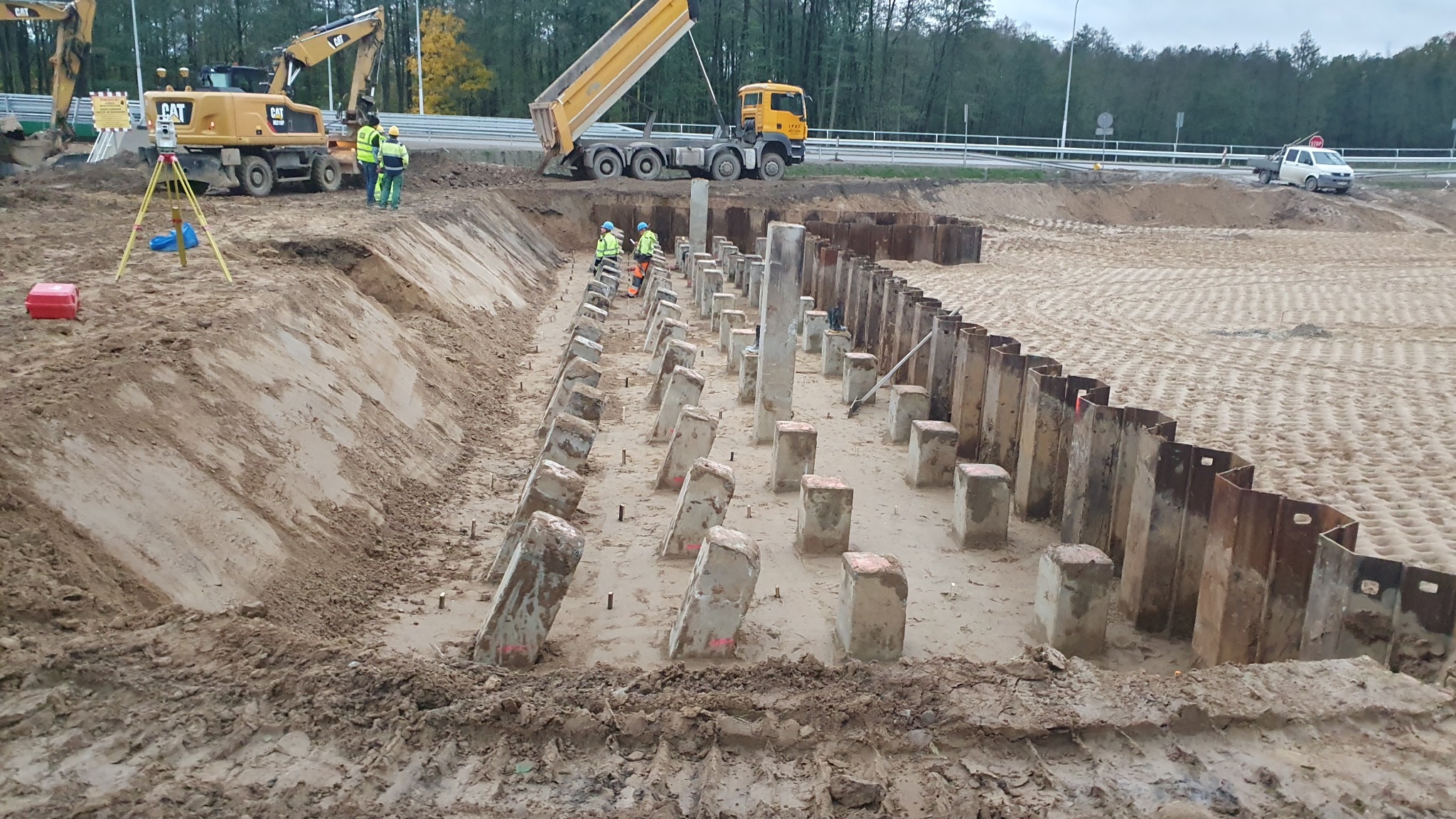 Prace ziemne z użyciem ciężkiego sprzętu: koparka, wywrotka i palownica. Widoczne betonowe pale i stalowe ścianki szczelne. Prace geodezyjne w tle.