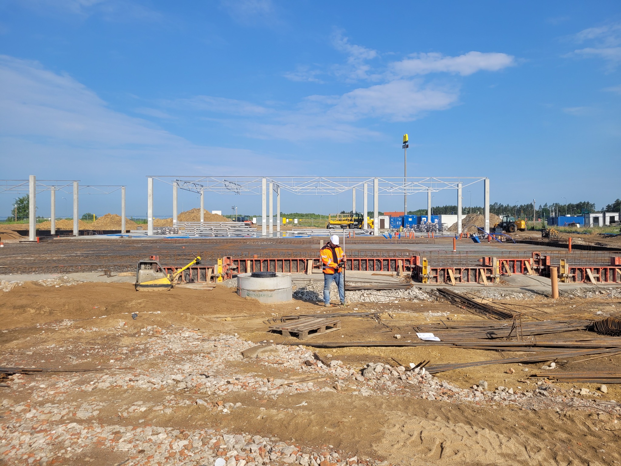 Pracownik w pomarańczowej kamizelce mierzy teren budowy zbrojenia pod budynek. Widoczne elementy konstrukcji i sprzęt budowlany w słoneczny dzień.