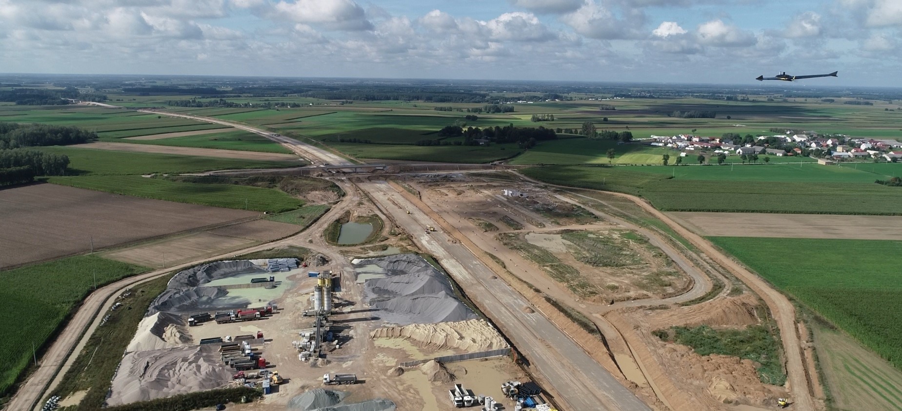 Szeroki kadr z drona na budowę drogi ekspresowej, z widocznymi maszynami budowlanymi, hałdami materiałów i okolicznymi polami. W tle widoczna wieś.
