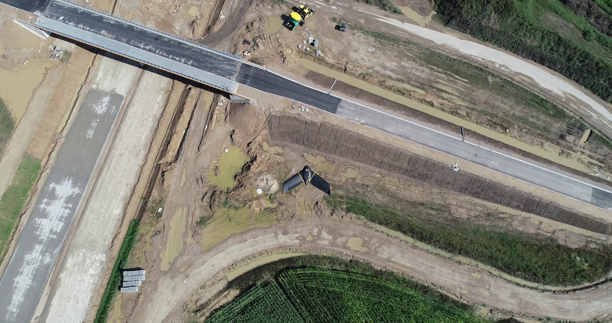 Zdjęcie z drona: budowa drogi z mostem, widoczne maszyny, teren budowy i fragmenty zieleni. Ujęcie z góry, ukazujące postęp prac.