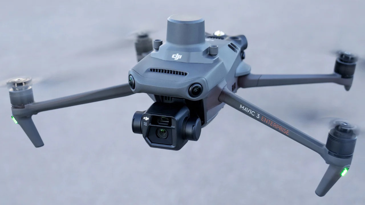 Szary dron DJI Mavic 3 Enterprise z kamerą, widoczne obracające się śmigła i zielone światła na końcach ramion, używany do pomiarów.