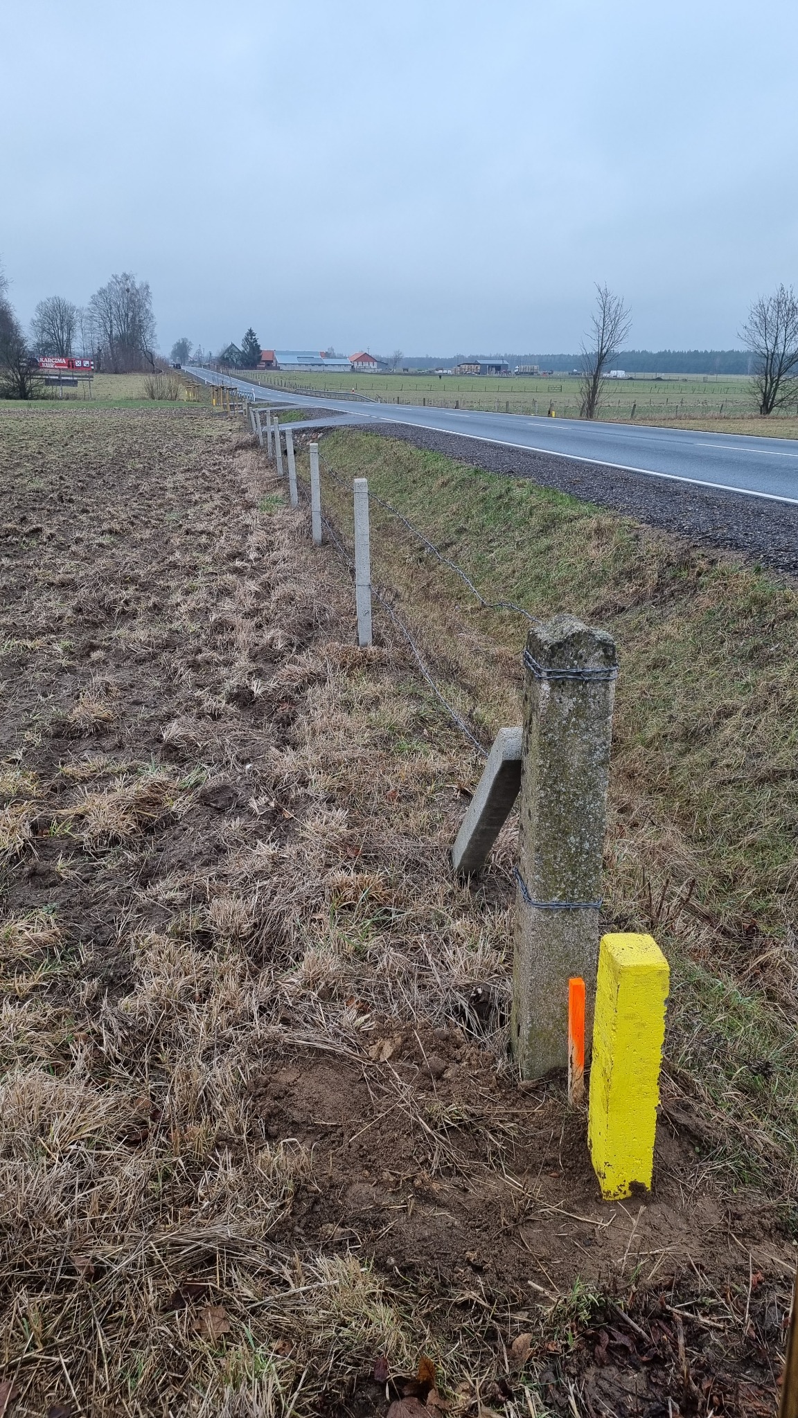 Granica działki wyznaczona słupkami z betonu i pomalowanymi znacznikami w jaskrawych kolorach, z widoczną drogą i polami w tle.