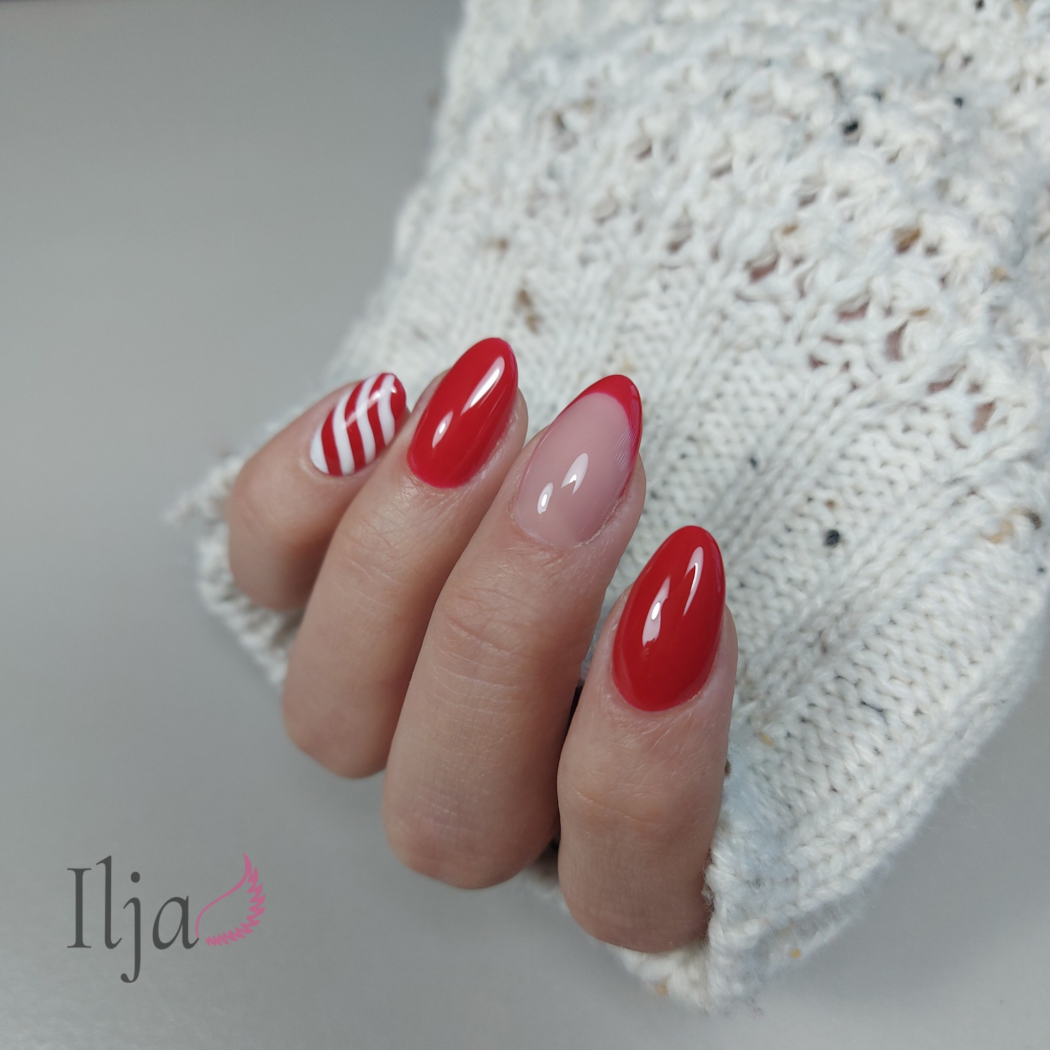 Dłoń w kremowym swetrze prezentuje stylizację paznokci: trzy czerwone, jeden z czerwono-białymi paskami, jeden z czerwonym french manicure.