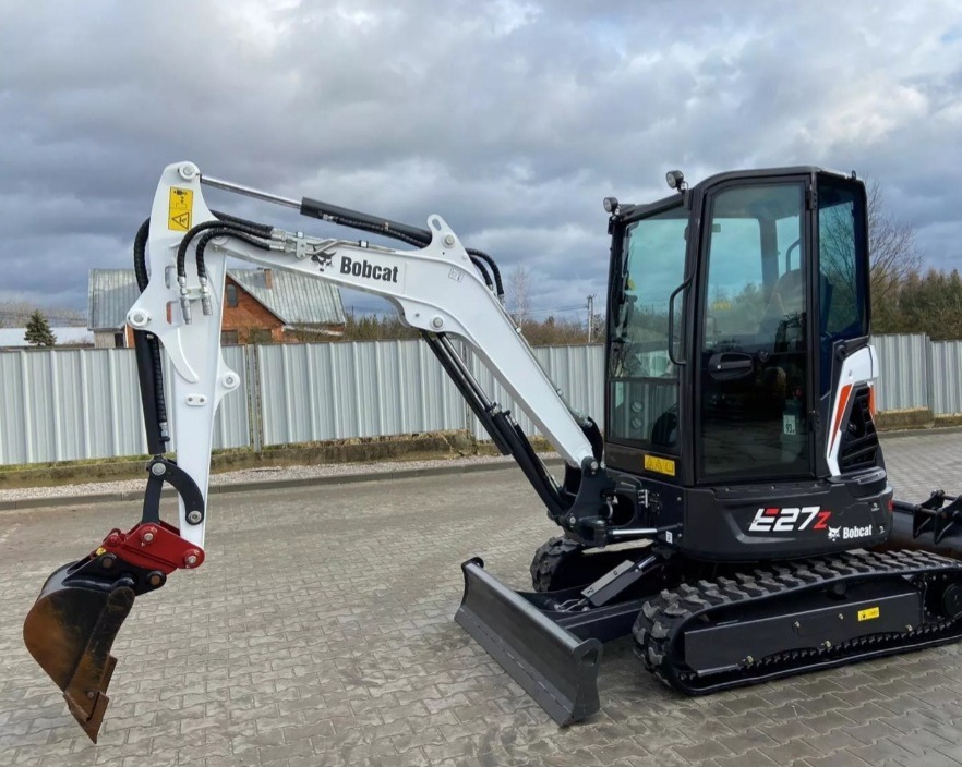 Kompaktowa, czarno-biała mini koparka Bobcat E27z z łyżką, zaparkowana na kostce brukowej na tle ogrodzenia z blachy falistej i pochmurnego nieba.