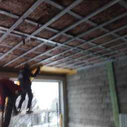 Patryk Franz - Montaż izolacji na poddaszu: pracownicy instalują żółte panele izolacyjne w metalowej konstrukcji sufitu na tle surowej ściany budynku w trakcie budowy.