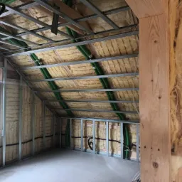 Poddasze w trakcie adaptacji: widoczne ocieplenie wełną mineralną, metalowy stelaż pod płyty gipsowe oraz zielone rury wentylacyjne pod stropem, okno z widokiem na zewnątrz.