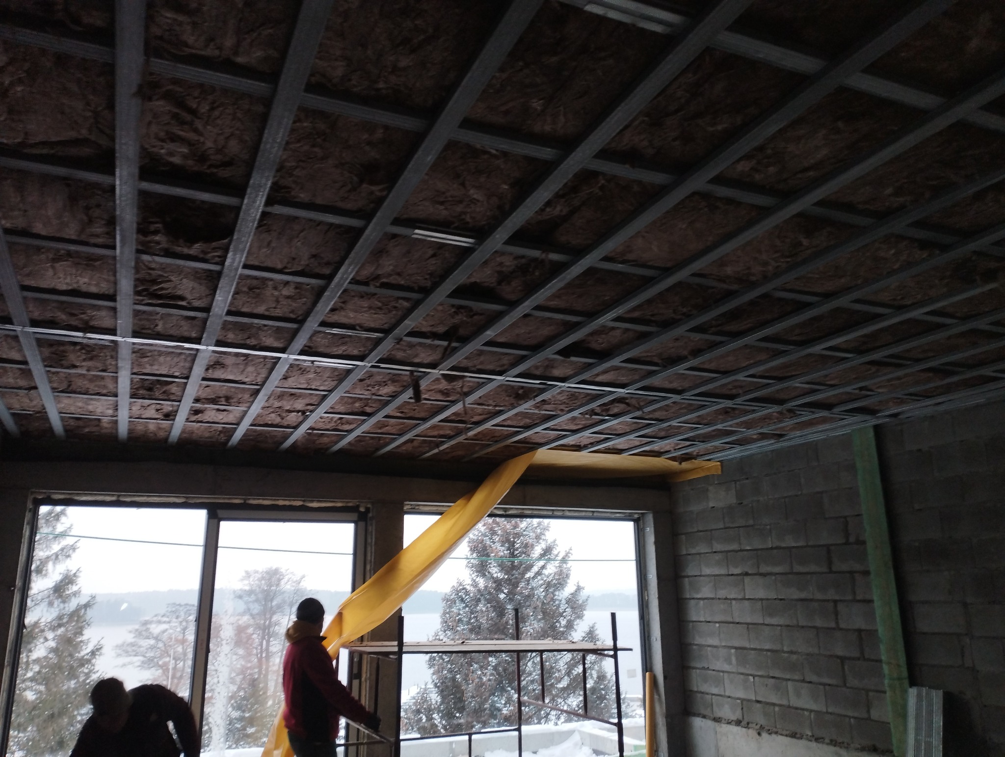 Montaż izolacji na poddaszu: widok na metalową konstrukcję sufitu z wypełnieniem z brązowej wełny mineralnej, częściowo osłoniętej żółtą folią, na tle okna z widokiem na zimowy krajobraz i jezioro,...