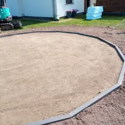 Przygotowany teren pod budowę, otoczony szarymi obrzeżami, z widocznym minikoparką w tle i stosem styropianu obok domu.