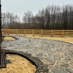 Ścieżka z nieregularnych kamiennych płyt, obrzeża z tworzywa, drewniane ogrodzenie w tle, przygotowane podłoże z piasku i żwiru.
