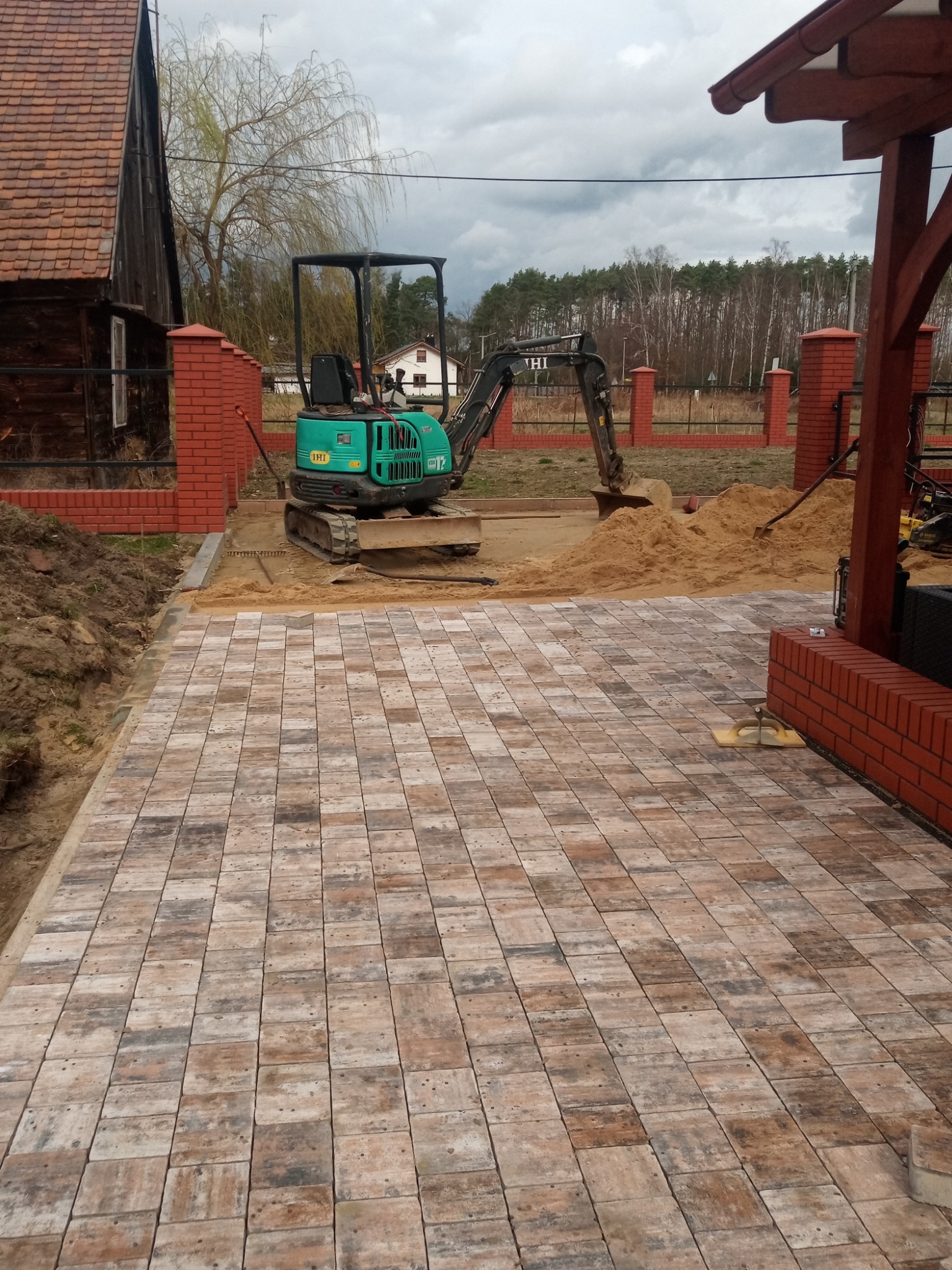 Ułożona kostka brukowa w odcieniach brązu i szarości, z widocznym mini-koparką przygotowującą teren pod dalsze prace, ceglane słupki ogrodzeniowe w tle i częściowo wykopana ziemia po lewej stronie