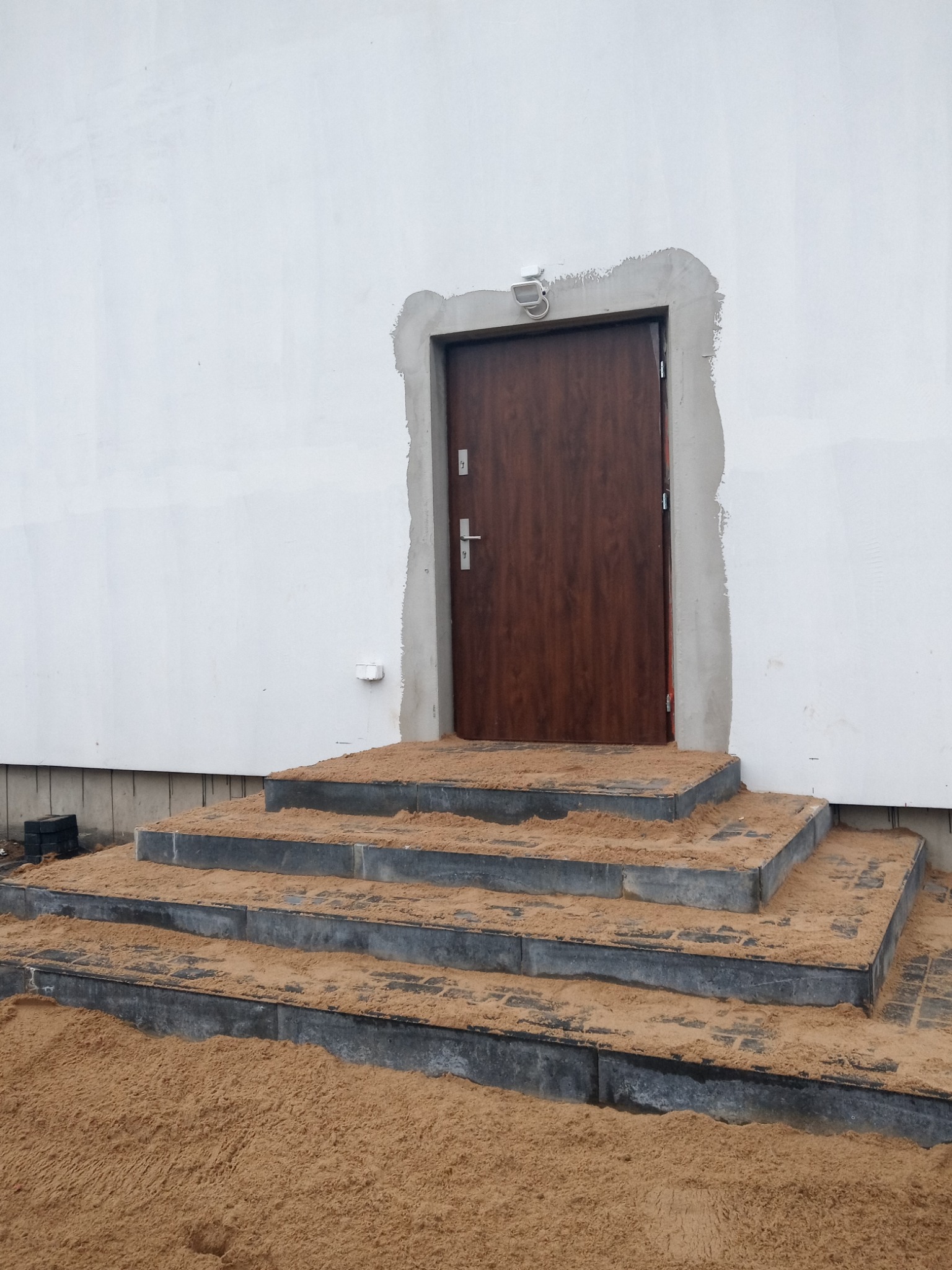 Wejście do budynku z drewnianymi drzwiami i schodami wyłożonymi szarą kostką brukową, częściowo zasypane piaskiem, ściana budynku biała, oświetlenie zewnętrzne nad drzwiami.