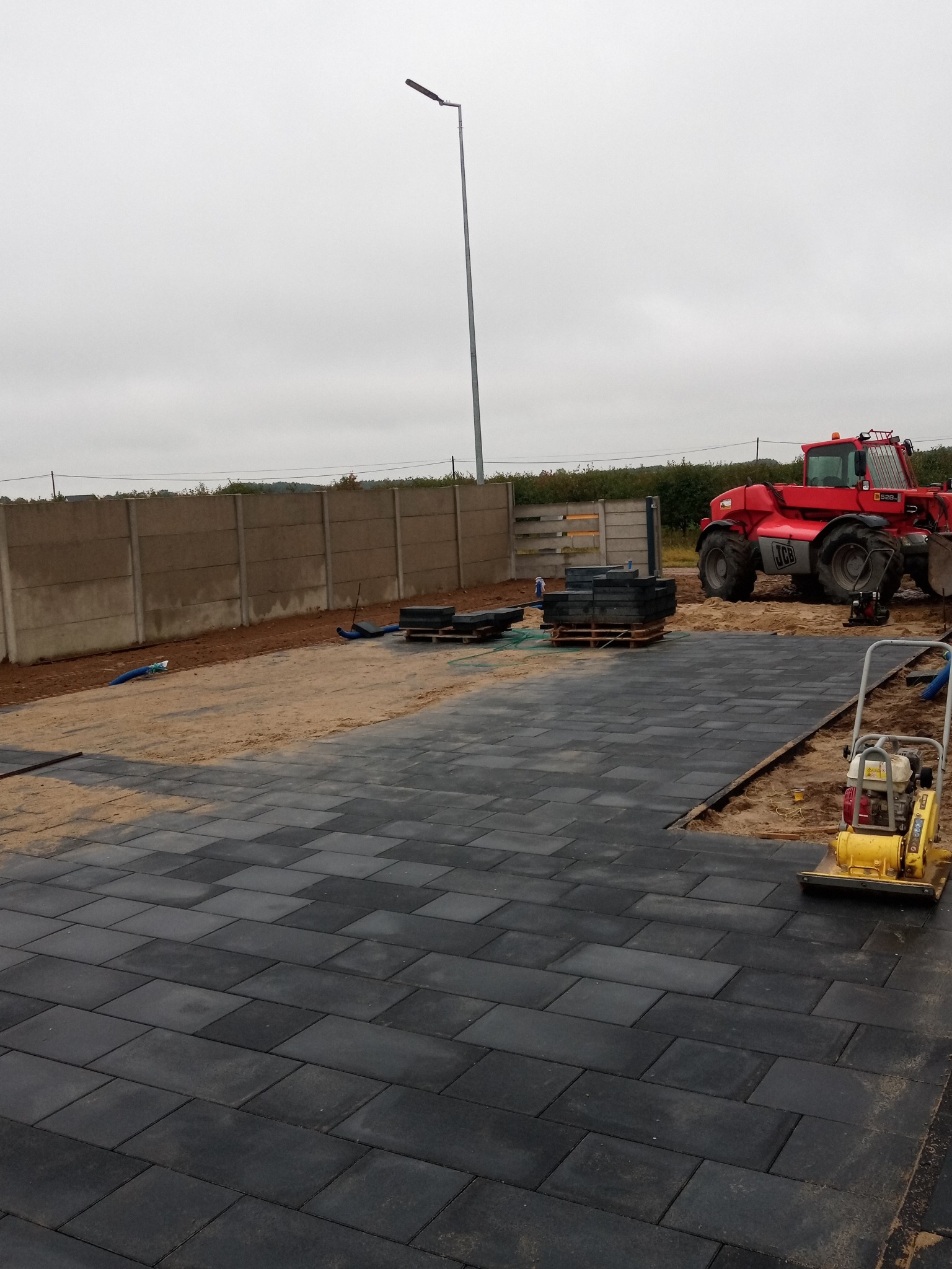 Częściowo ułożona, ciemnoszara kostka brukowa w trakcie prac, widoczna zagęszczarka wibracyjna, paleta z kostką, czerwona ładowarka teleskopowa JCB w tle i betonowe ogrodzenie panelowe.