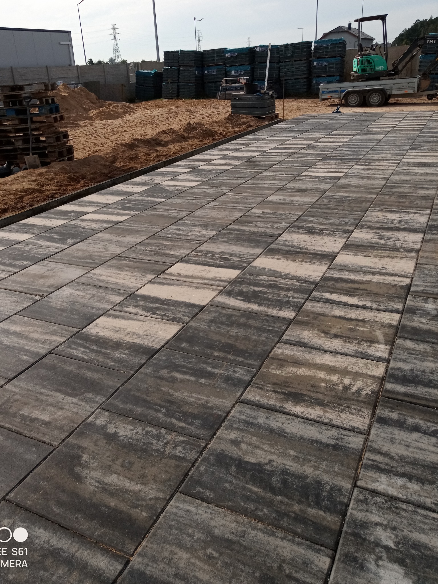 Ułożona kostka brukowa w odcieniach szarości i beżu, z widocznym przygotowanym podłożem z piasku, paletami i mini koparką w tle.