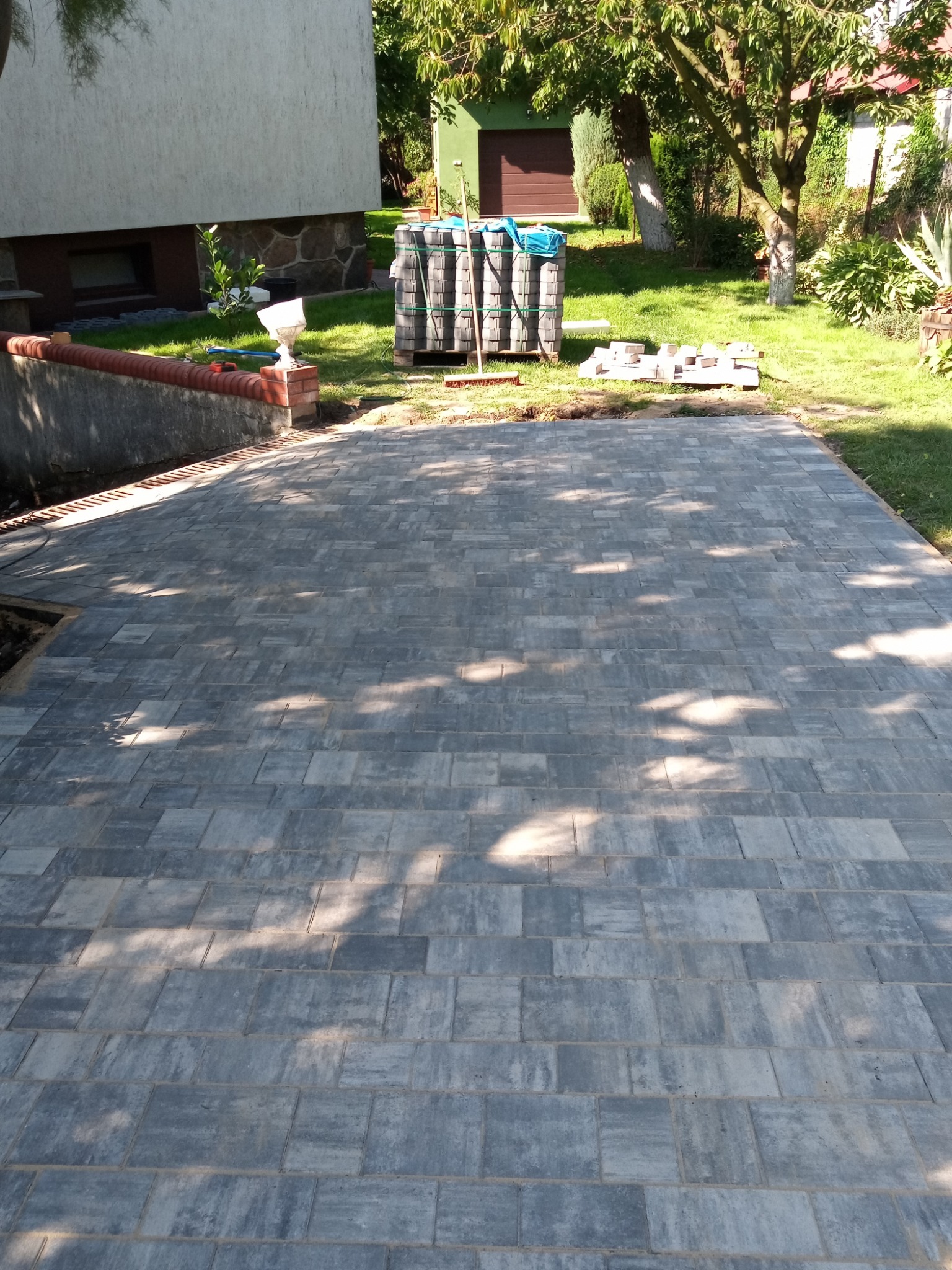Ułożona szara kostka brukowa na posesji, widoczne palety z kostką w tle oraz fragment budynku z oświetleniem zewnętrznym.